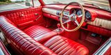Red leather interior of a classic convertible car, with wooden steering wheel and dashboard gauges.