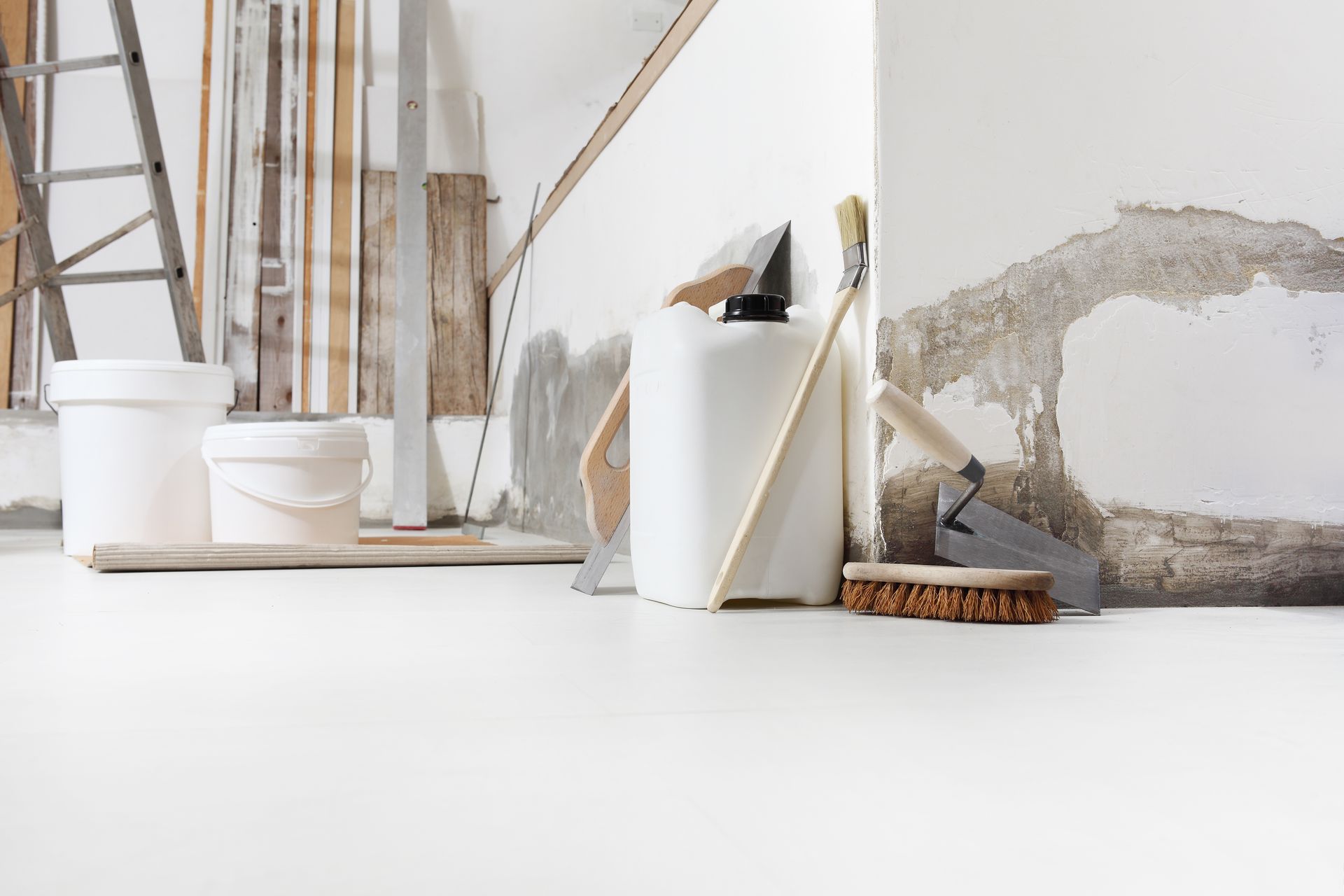 White buckets, tools, and a plastic container sit on a clean floor against a wall undergoing renovation.