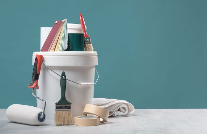 A white bucket filled with painting supplies, including rollers, brushes, masking tape, and cloths against a blue wall.
