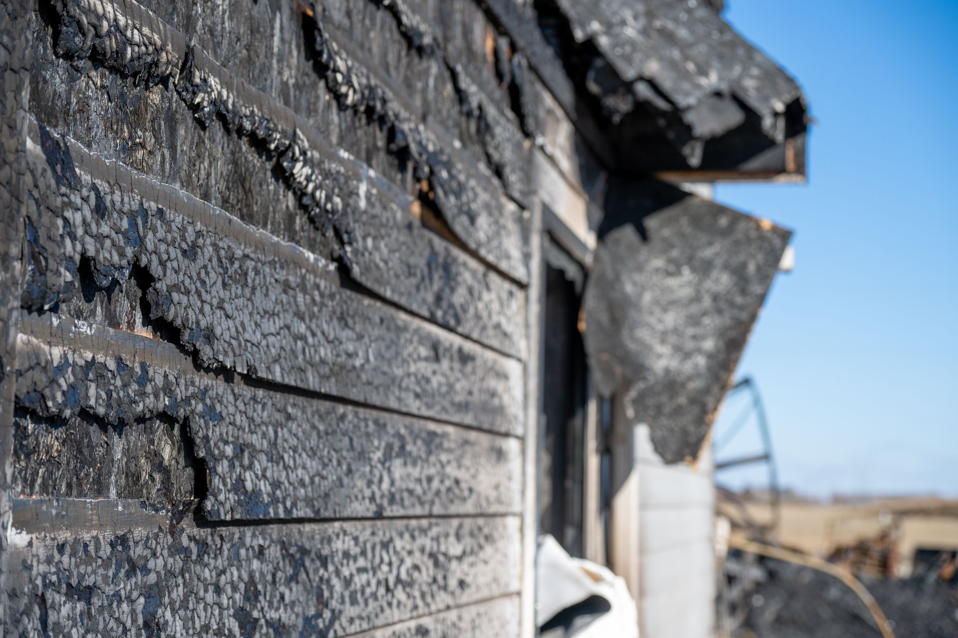 Close-up of charred house siding with heavy fire damage under clear blue sky. Close-up of charred house siding with heavy fire damage under clear blue sky.