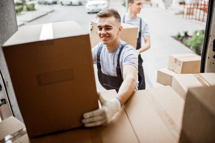Movers loading cardboard boxes into a van; one smiling, wearing gloves.
