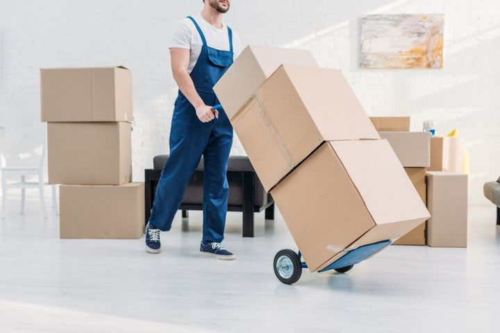 Man in blue overalls moving stacked cardboard boxes with a dolly in a room.