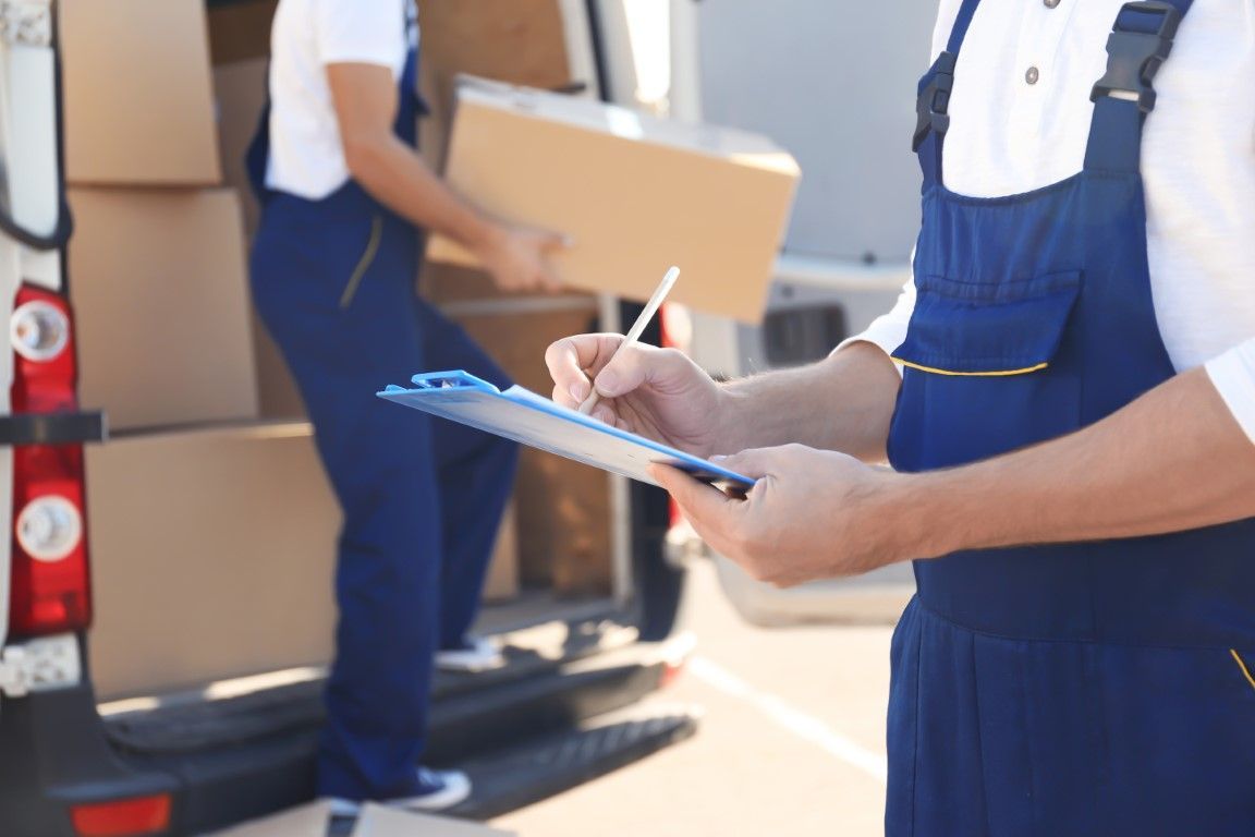 Two movers loading boxes into a van. One writes on a clipboard; the other carries a box.