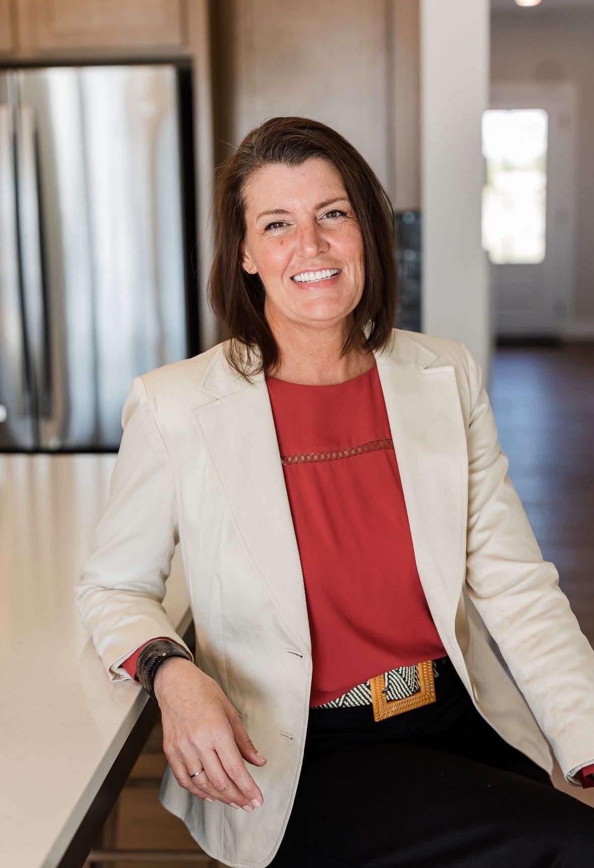 Woman in beige blazer and red top smiles, leaning on a white kitchen counter.