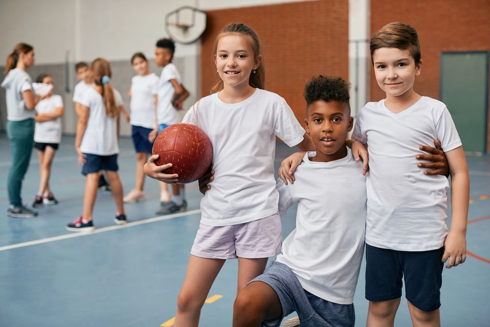 Tre bambini in palestra: una bambina che tiene in mano una palla e due bambini che si abbracciano, sorridenti. Altri bambini sullo sfondo.