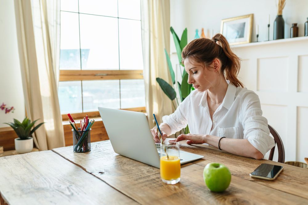 Donna che scrive a un tavolo di legno con un computer portatile, del succo di frutta e una mela. Stanza luminosa, luce naturale.