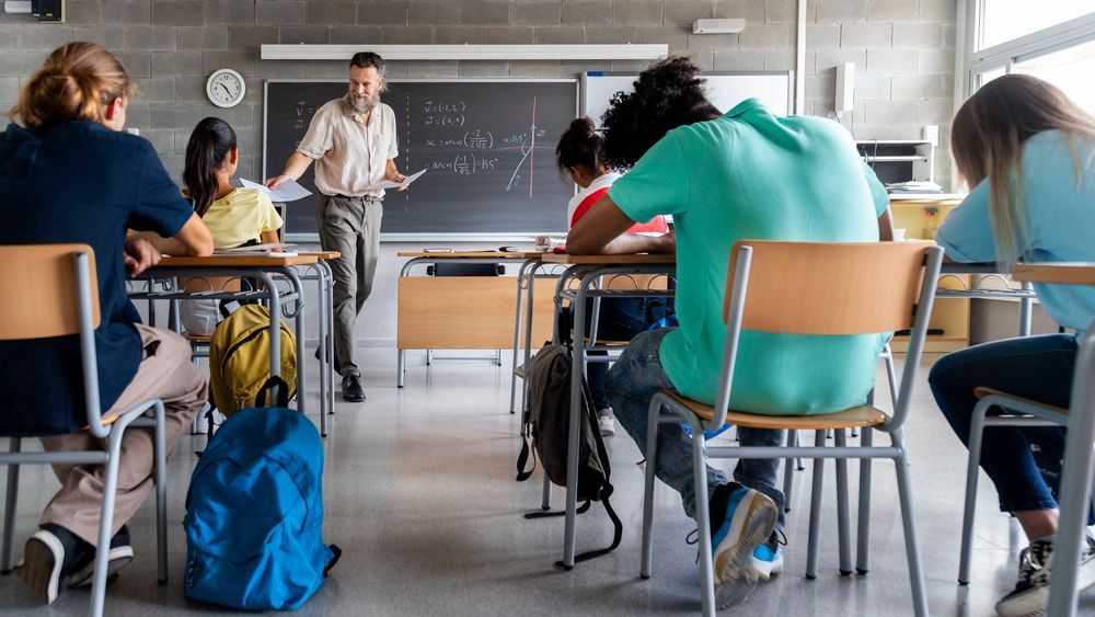 Insegnante in piedi alla lavagna che tiene lezione agli studenti in classe. Studenti seduti ai banchi che guardano gli appunti.