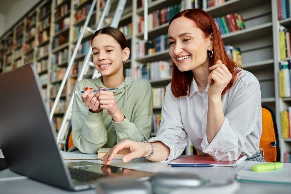 Due persone sorridono davanti a un computer portatile in una biblioteca. Una indica lo schermo e l'altra tiene una penna.