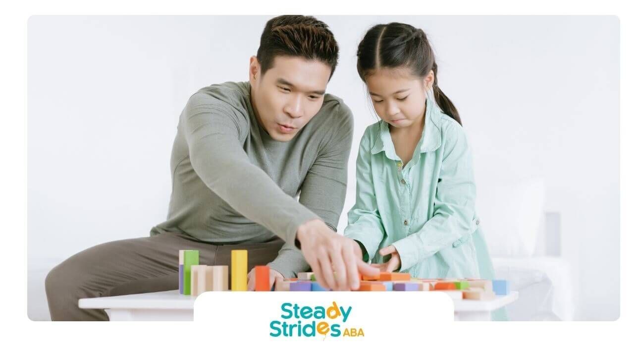 Therapist helping autistic girl build colorful wooden blocks at a small table during learning play.