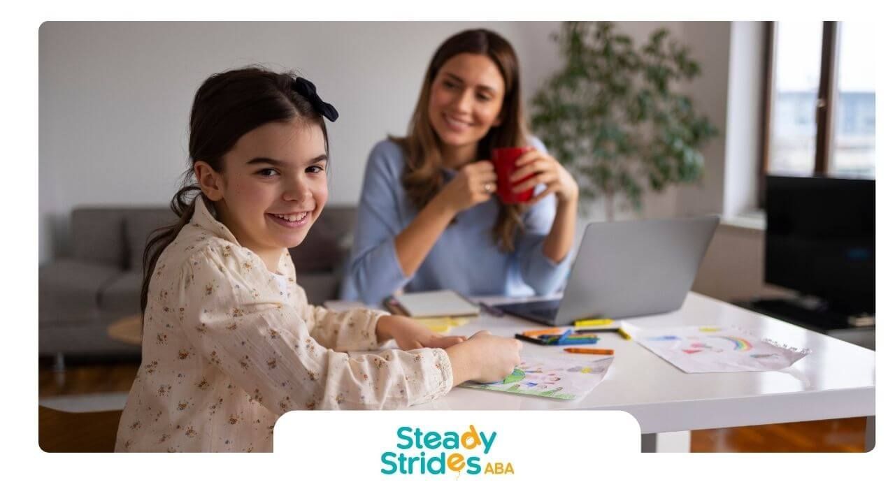 Autistic girl smiles while sitting at a table with therapist in the background during therapy.