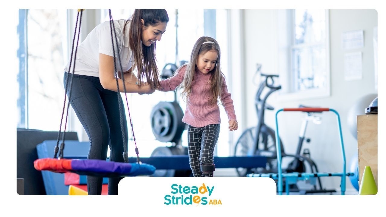 Woman assisting a child walking in a therapy room, near a swing and exercise equipment.