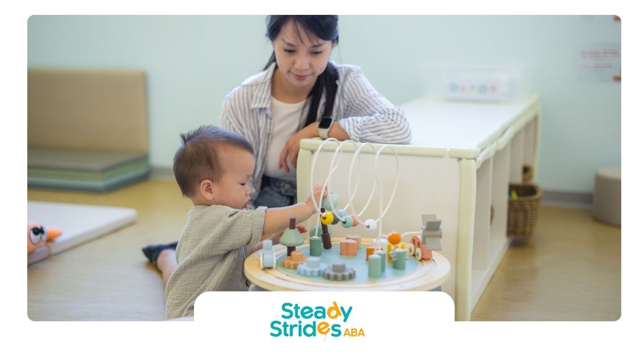 Baby playing with a wooden activity toy, supervised by a person, in a light-filled room.