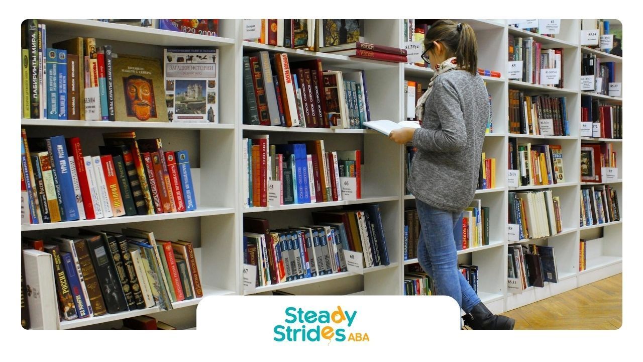 An ABA therapist reading an ABA therapy book in library aisle surrounded by shelves in Texas.
