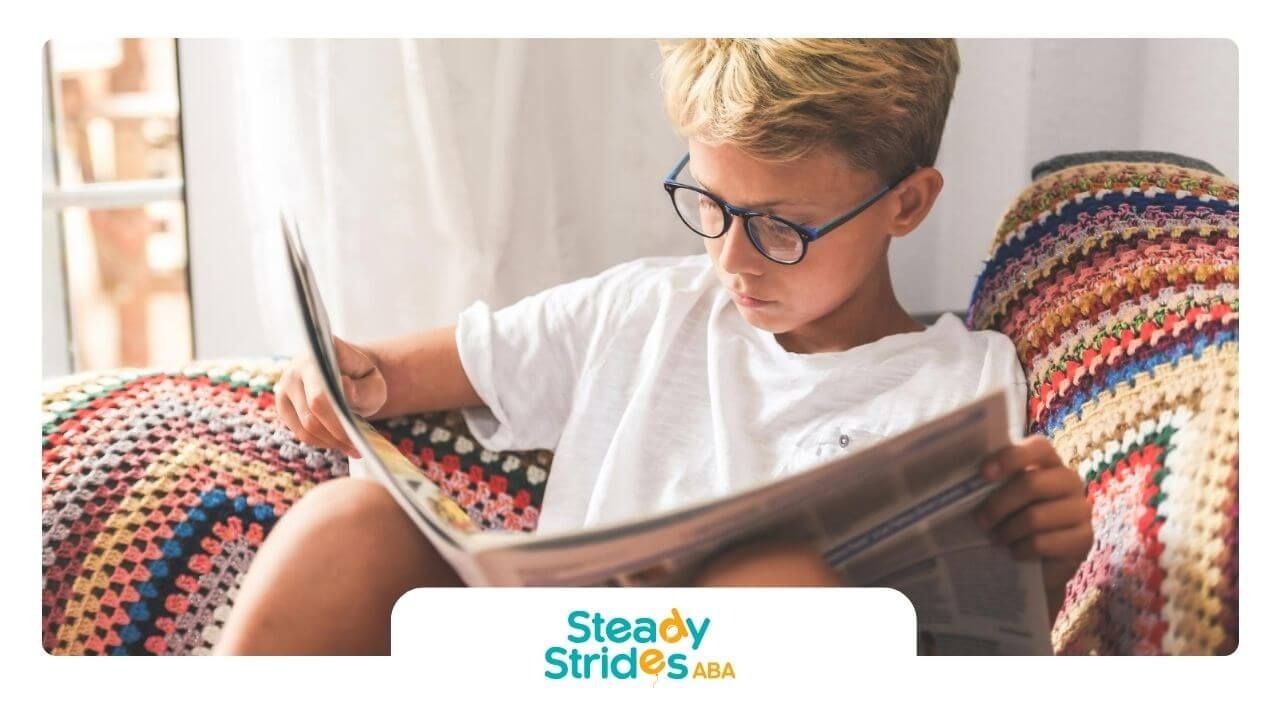Smiling boy with autism sitting at a table reading a book indoors.