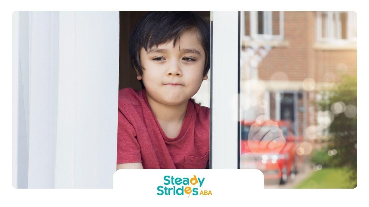 A boy with autism in red shirt looking out the window thoughtfully.