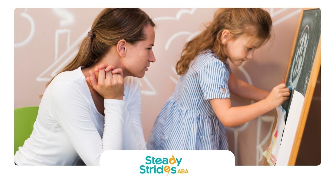A therapist observing a autistic girl drawing on a chalkboard during a supportive learning activity.