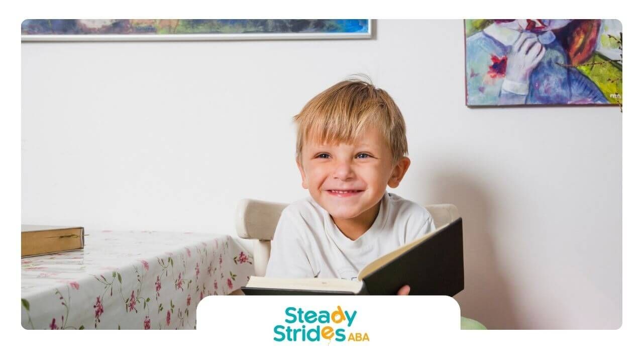 Smiling boy with autism sitting at a table reading a book indoors.