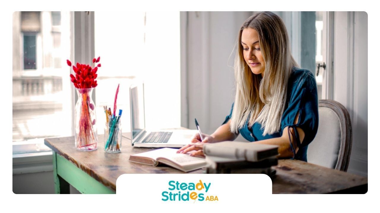 A BCBA aspirant writing in a notebook while studying on her laptop at a desk by the window in Texas.