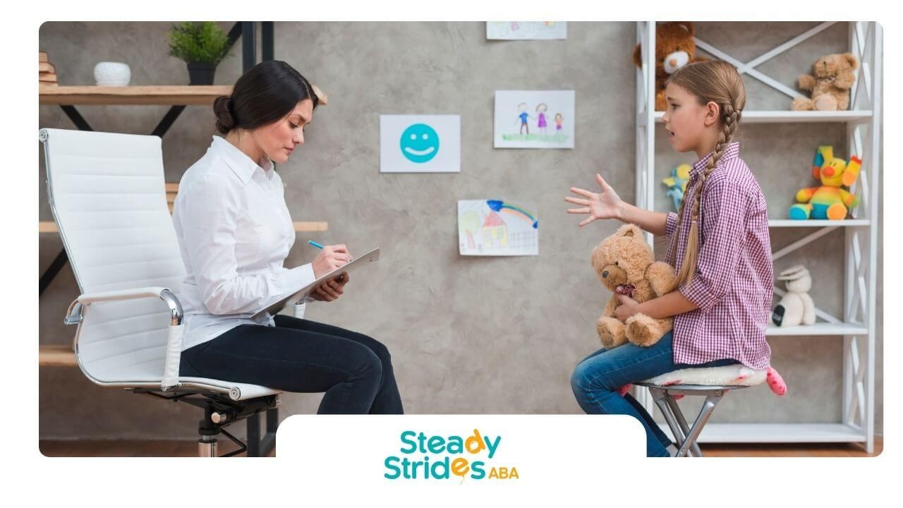 Therapist taking notes while a n autistic girl speaks & holds a teddy bear during session.