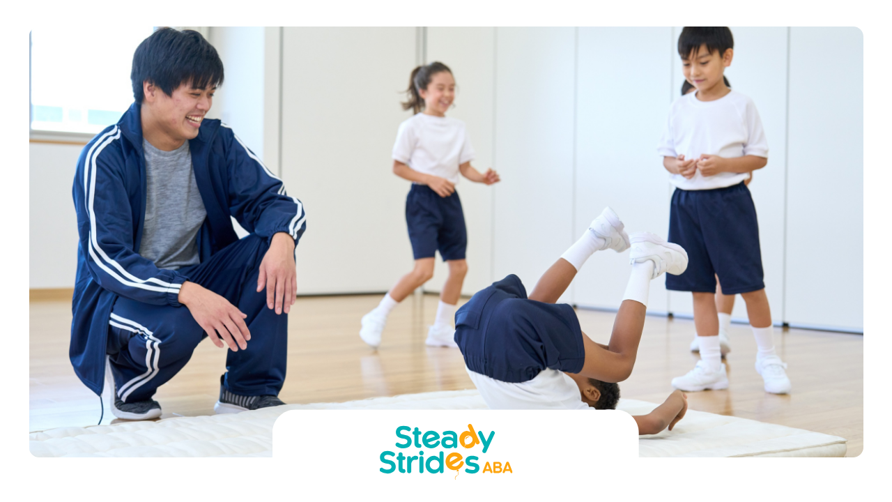 A man watches a child perform a forward roll as two others play, all in a gym.
