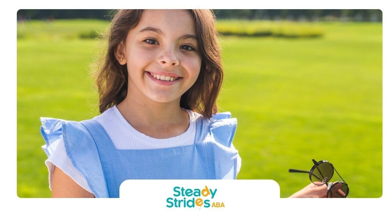 Smiling autistic girl holding sunglasses outdoors in a sunny park, showing confidence and happiness.