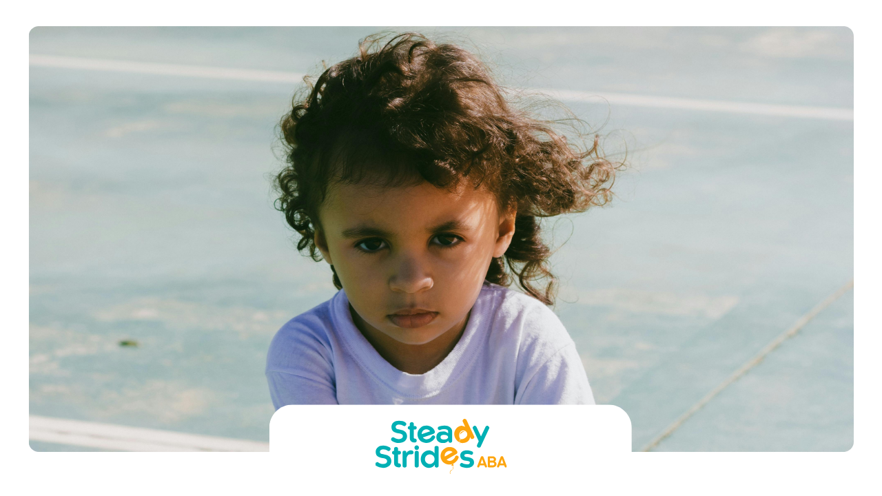 Child with curly hair in a white shirt, looking at the camera. Soft-focus background.