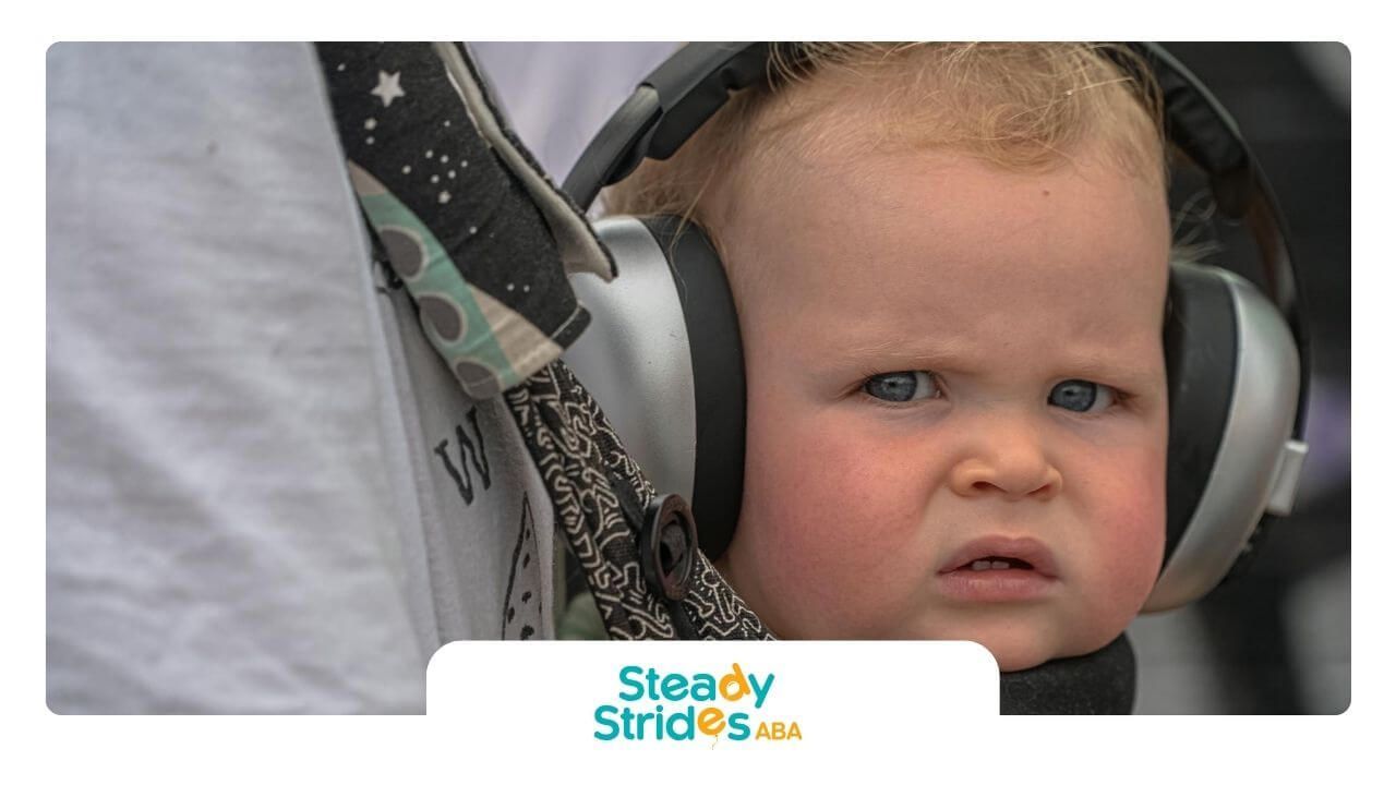 Autistic child with headphones looking confused in a carrier, possibly overwhelmed by sounds.