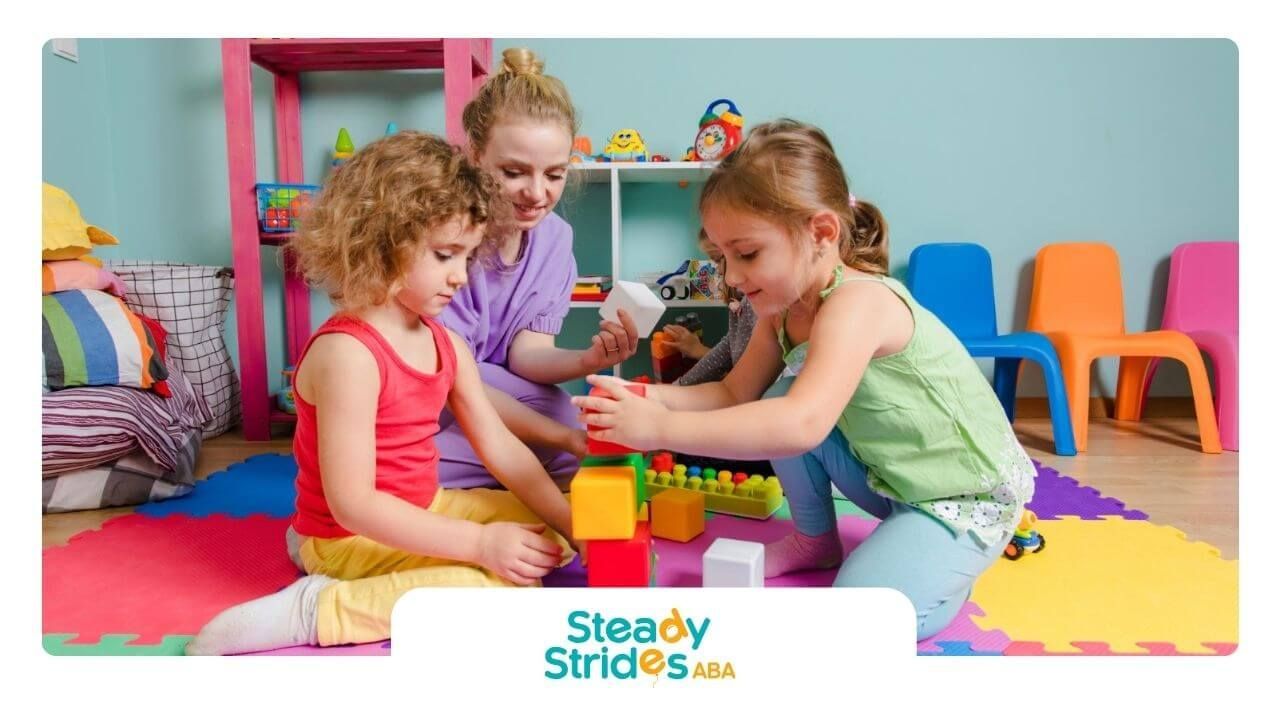 Two girls with autism play with blocks while a BCBA helps during ABA therapy.