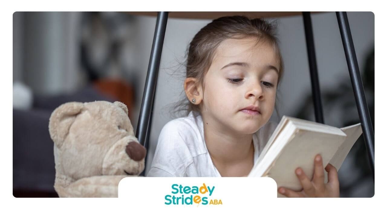 A little girl with autism is sitting in a chair reading a book next to a teddy bear.