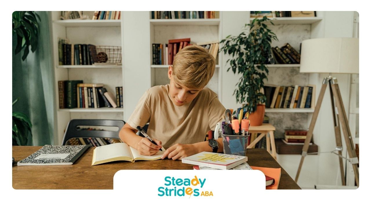 An autistic boy writing in a notebook at a desk surrounded by books during ABA therapy in Texas.