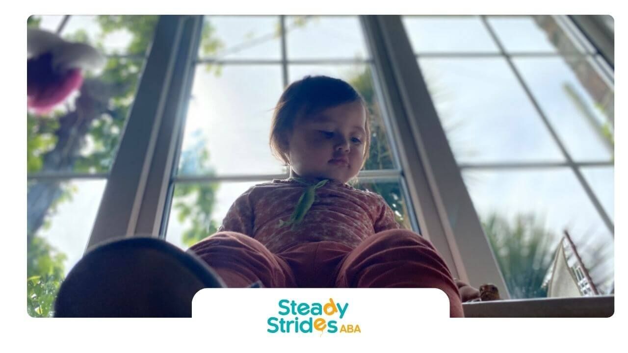 Autistic toddler sits on a windowsill looking down at a toy, exploring balance and spatial awareness