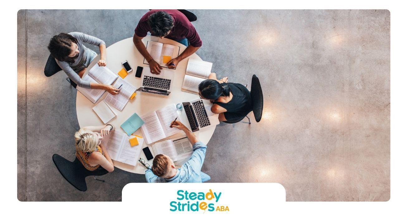 Overhead shot of five people studying around a round table with books, laptops, and phones.
