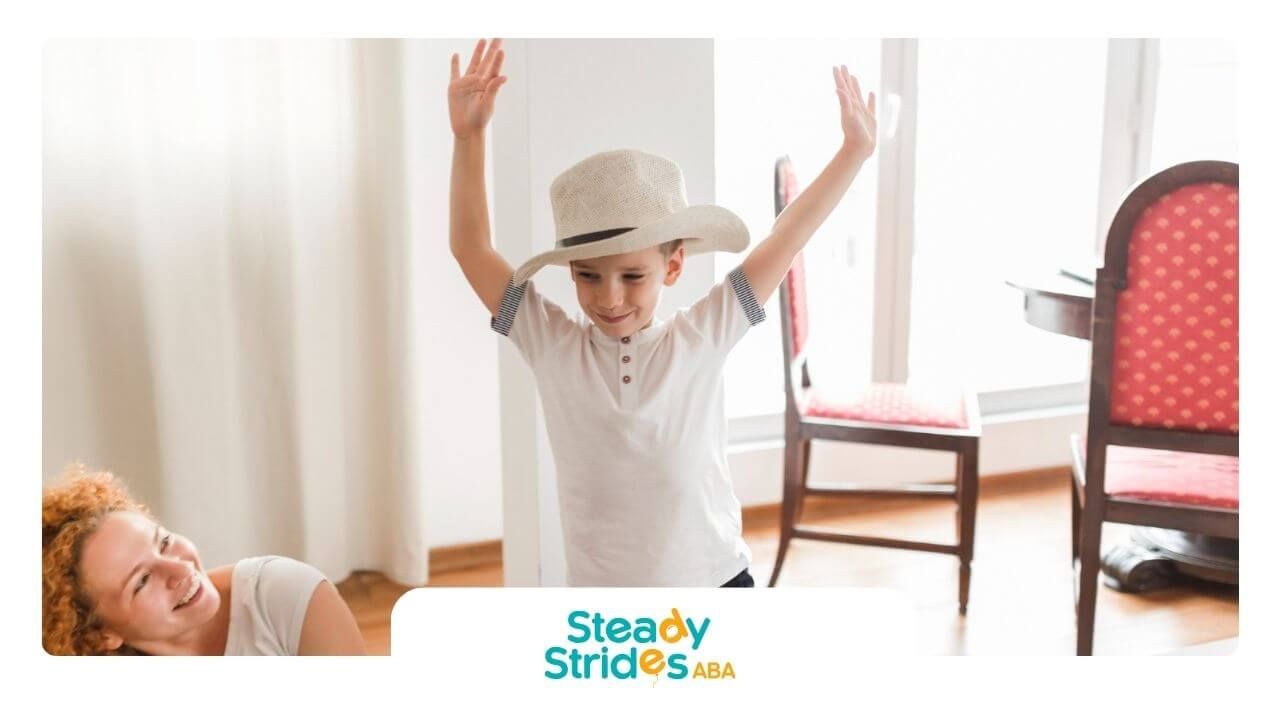 A boy with autism in a cowboy hat raises his arms while an ABA therapist smiles.
