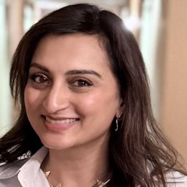 Person in graduation cap and gown, smiling slightly. Gold and black regalia, dark background.