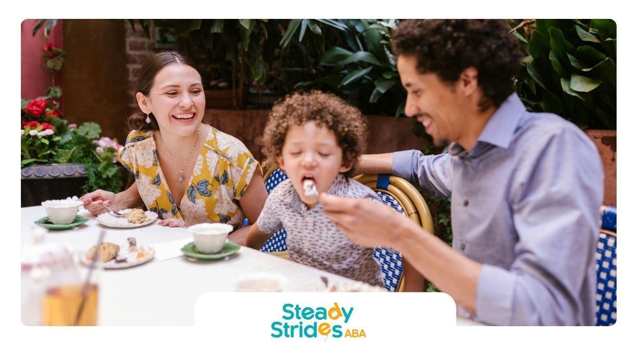 Parents laughing while feeding young autistic child dessert at restaurant during ABA therapy in TX.