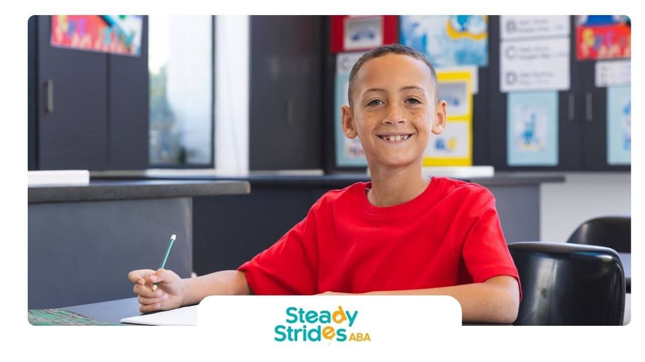 Autistic boy in a red shirt is sitting at a desk in a classroom, smiling and preparing to write.
