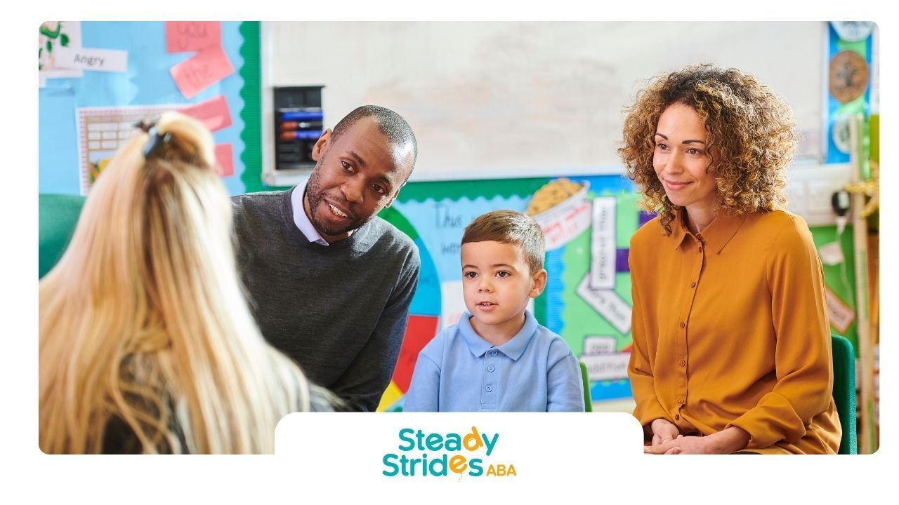 Classroom meeting: Boy, teacher, and two adults, smiling. Whiteboard and posters in background.