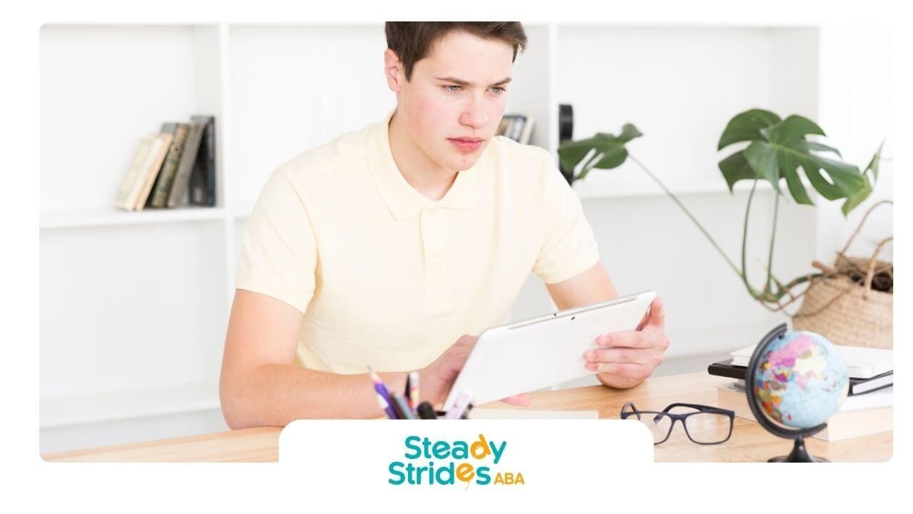 Male ABA therapist studying at desk with tablet, notebook, globe, and books in bright office.