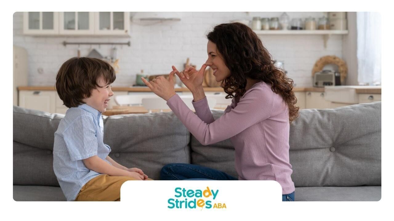 An ABA therapist and an autistic boy smiling and using hand gestures while sitting on a couch.