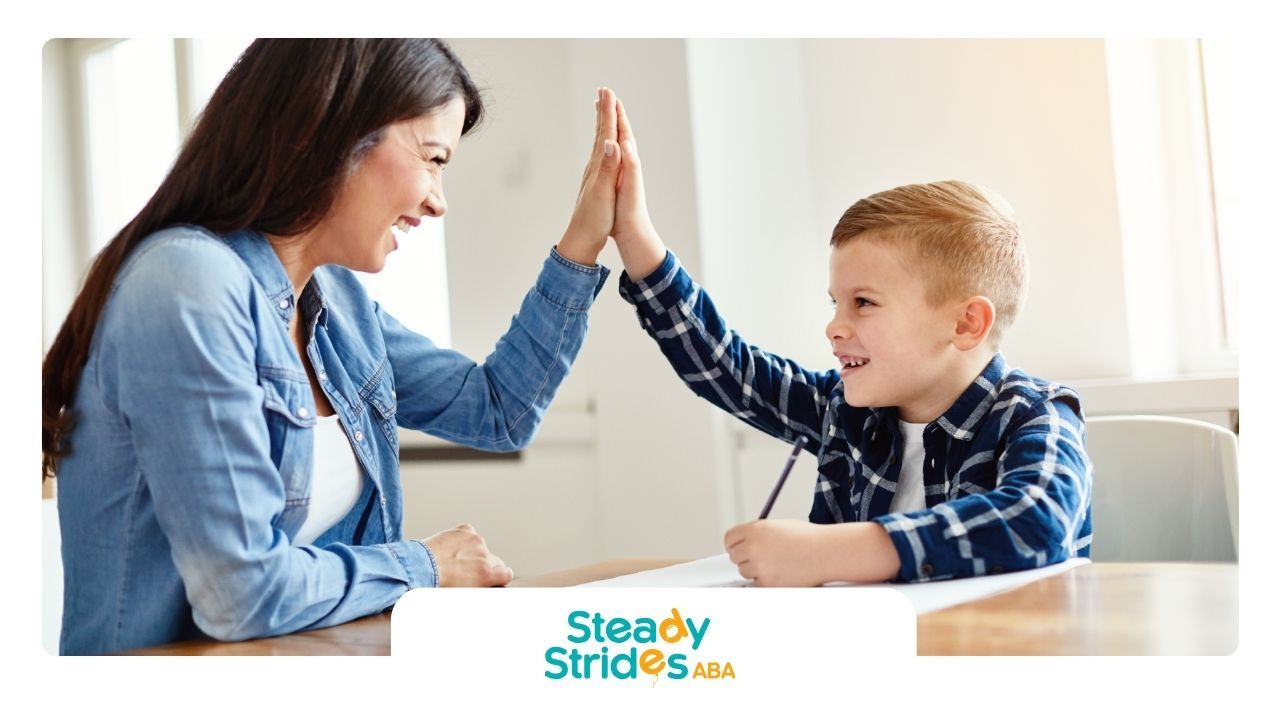 Woman high-fives a smiling boy at a table; he wears a plaid shirt. Light-filled room.