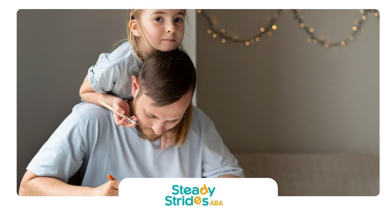 Therapist draws with markers at a table while an autistic girl leans on his shoulders during therapy