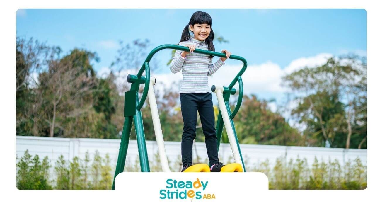 Autistic girl is playing on a playground swing, holding the structure with both hands while smiling.