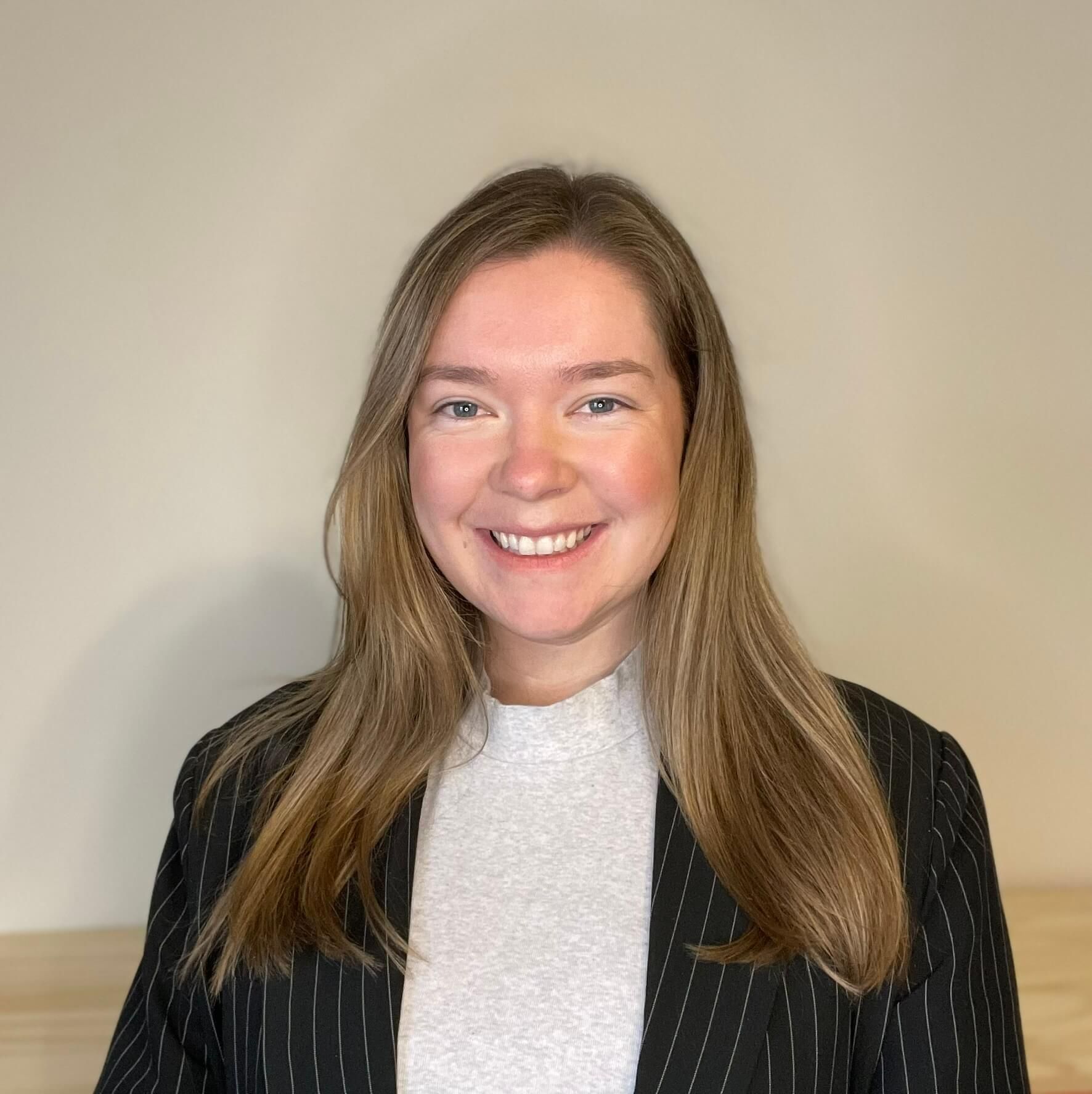 Person in graduation cap and gown, smiling slightly. Gold and black regalia, dark background.