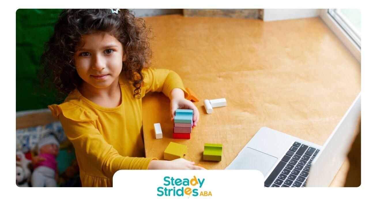Autistic girl stacking colorful blocks beside a laptop while playing and learning at a wooden desk.