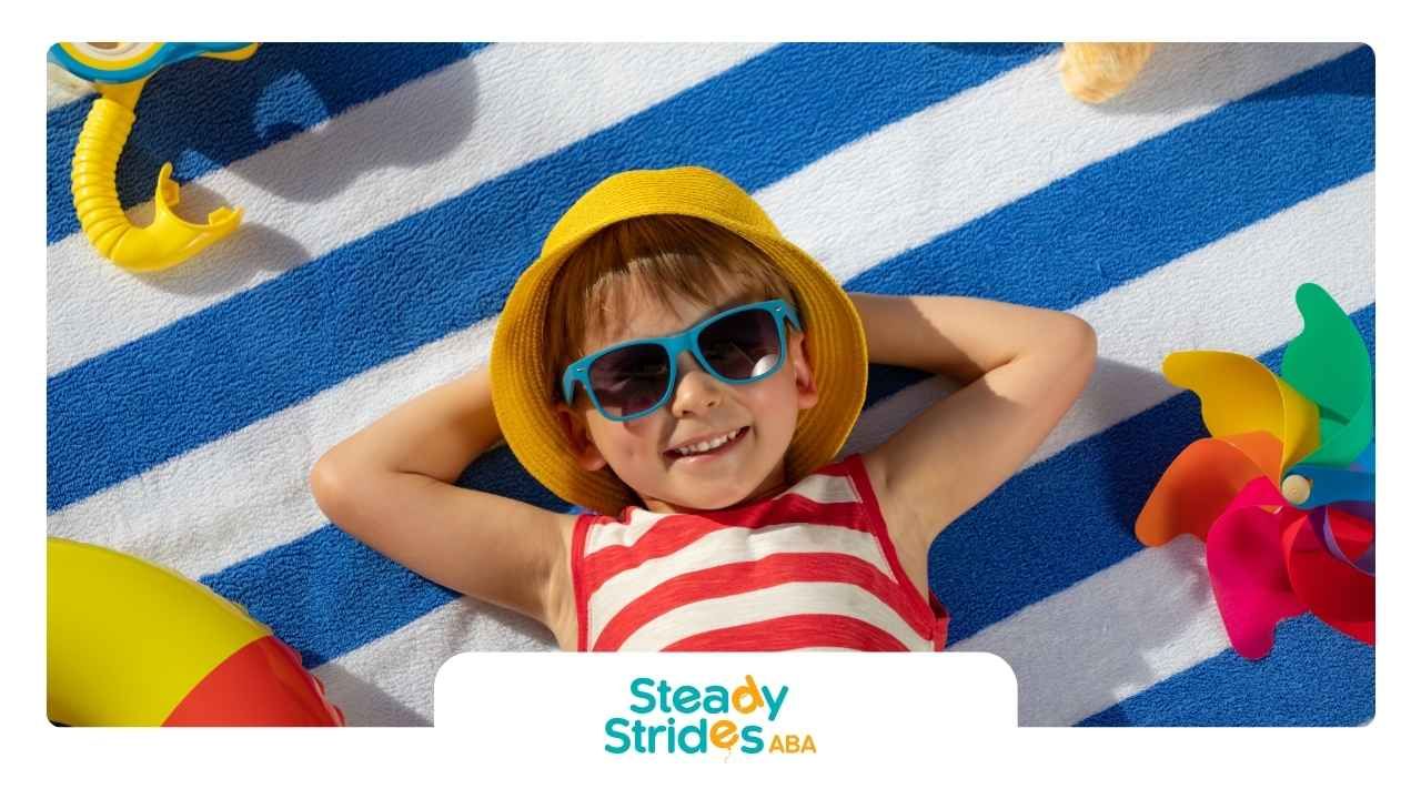A joyful child in sunglasses and a yellow hat relaxes on a blue-striped towel.