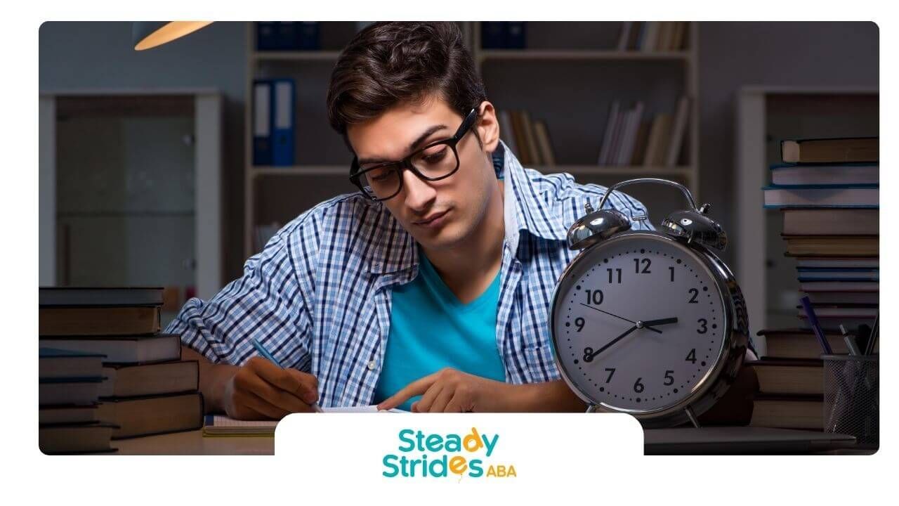 A therapist wearing a graduation cap sits at a desk with books, a clock, and study materials
