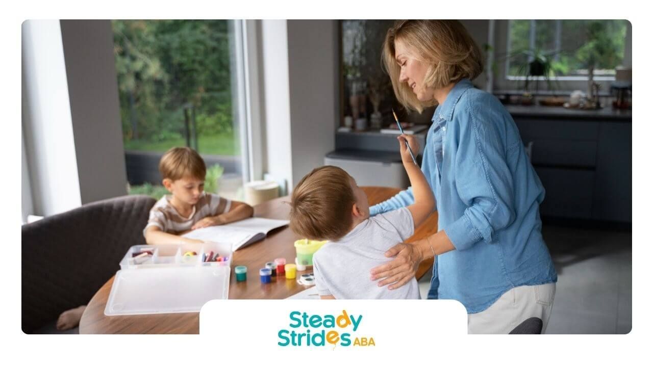 A BCBA is standing next to two children sitting at a table in a living room.