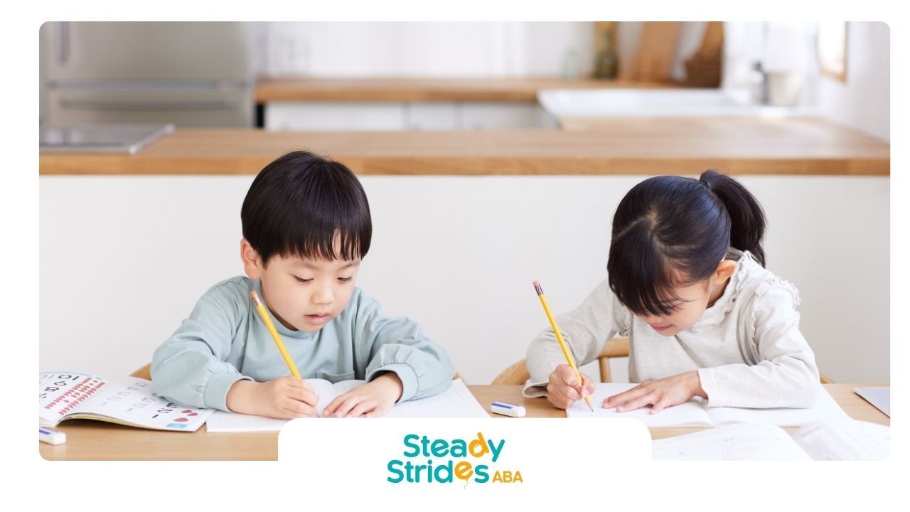 Two children sitting at a table writing with pencils in a well-lit room.