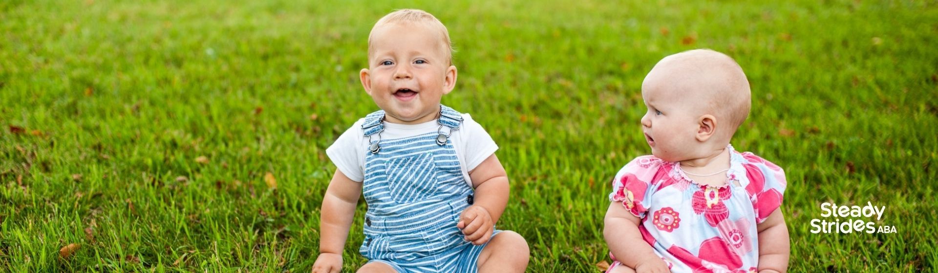 Two babies sitting in green grass, one smiling, the other looking to the side.