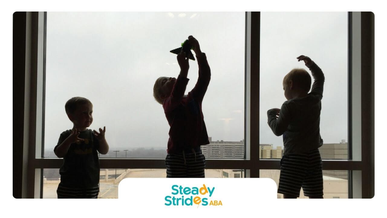 Three kids in striped shorts pose on windowsill with toy airplane in front of large window